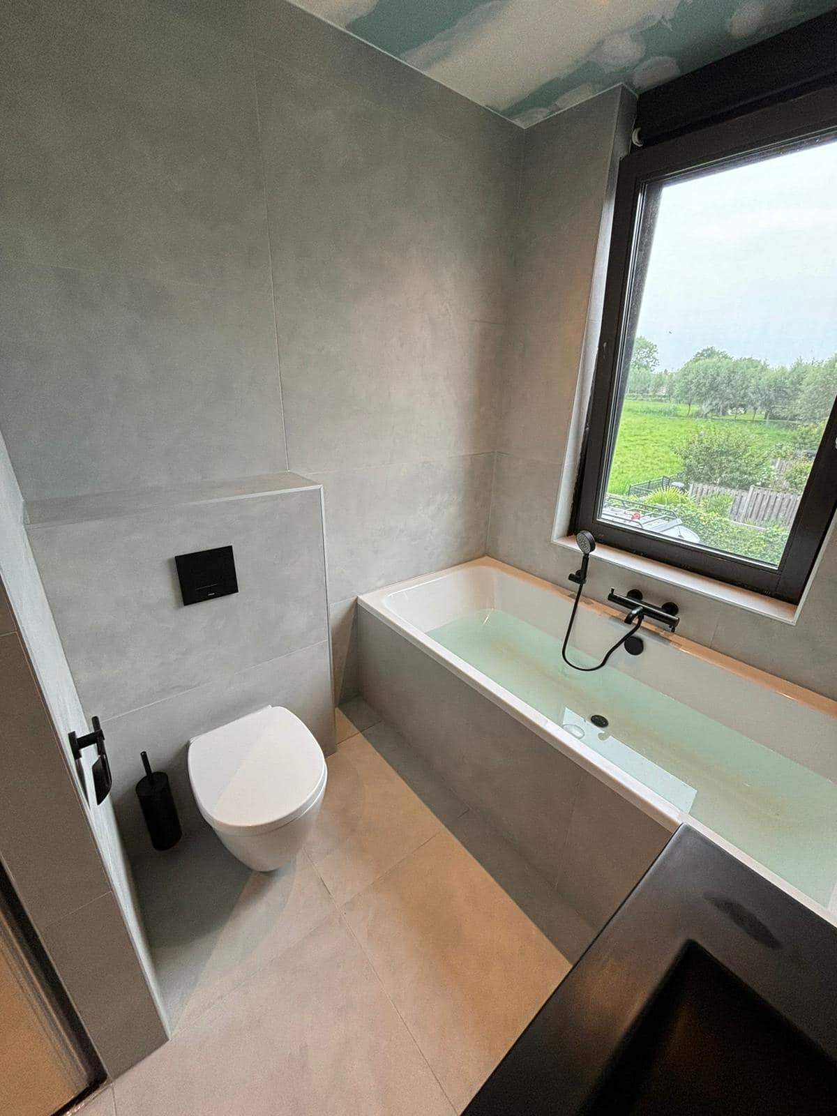 Badkamer met ligbad: Ligbad onder het raam, wandcloset en grote grijze tegels.